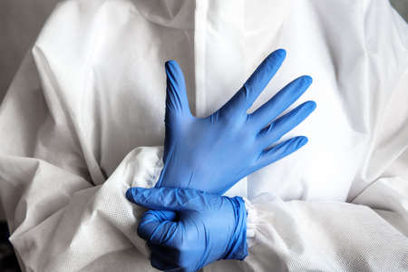 Doctor or nurse in white medical PPE suit puts on protective gloves, physician in hospital close-up.
