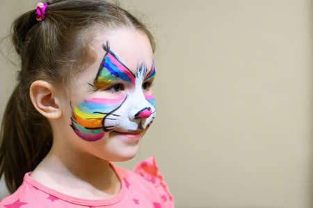 Kid with face painting of kitty, cute little girl with painted mask on face of rainbow cat. Portrait of pretty child with beautiful makeup and copy space background, unique facial body art for party.の写真素材