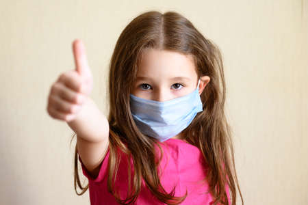 Kid in face mask for protection to corona virus shows thumb up, portrait of pretty little girl wearing medical mask due to COVID-19. Cute child staying at home during COVID coronavirus pandemic.の写真素材