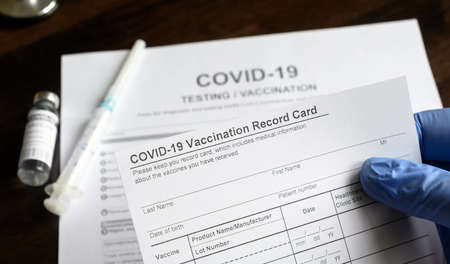 COVID-19 Vaccination Record Card in doctor hand, coronavirus test and vaccine medical form on desk in clinic. Concept of corona virus vaccination certificate, health passport and immunization.の写真素材