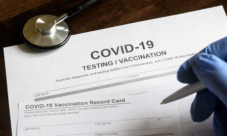 COVID-19 testing and vaccination forms on desk, doctor makes an entry in coronavirus vaccine record card, medical document in clinic. Concept of corona virus vaccination certificate and immunization.の写真素材