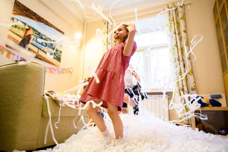 Happy kid on party, little girl having fun with confetti in playroom, paper show at home. Child plays, dances and laughs on birthday in room. Active celebration, expression, mess and disco concept.の写真素材