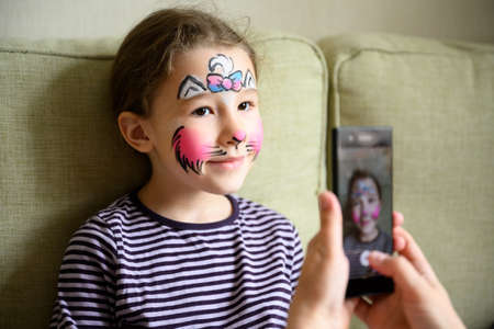 Kid with face painting of kitty, cute little girl with painted mask on face poses for photo. Happy...
