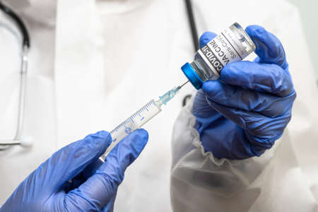 COVID-19 vaccine in doctor hands, physician in PPE suit holds syringe and vial with corona virus...