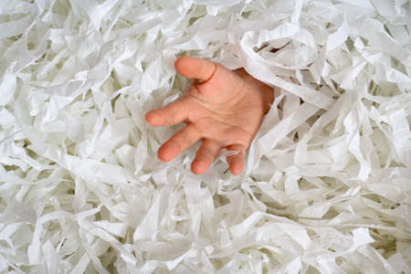 Hand reaches out from heap of shredded paper, man sinking in pile of white confetti. Somebody drowning in paper trash, garbage or box filler. Concept of paperwork, party, office work and shipping.の写真素材