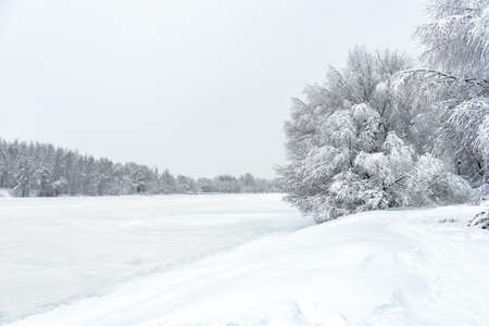 Winter forest landscape, snowy ice river during snowstorm for background. Scenic view of riverbank trees under snow. Concept of seasonal weather, Siberia, forecast, frozen white nature and climate.の写真素材