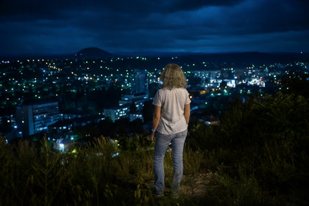 Panorama of Pyatigorsk at night, Russia. Young woman stands on city and sky background, person tourist looks at town lights from mountain top in summer. Theme of travel people, hiking and tourism.の写真素材