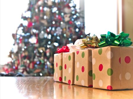 Four beautifully wrapped gift boxes on wood table with colorful bows, with decorated Christmas tree...