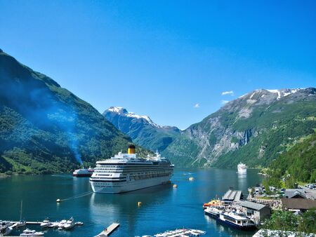 Spectacular view of Geiranger Fjord and mountains with snow, waterfalls, moored cruise ships and tender boats carrying tourists from ship to shore on a beautiful day with blue sky.の写真素材
