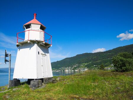 Close view of white lighthouse tower with red top along the shore of the Sognefjord in Norwegian on a sunny day, with mountains, blue sky and fluffy white clouds.の写真素材