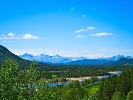 Beautiful farm valley landscape with a flowing river, snow capped mountains and green trees under a...