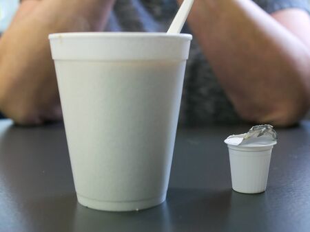 Styrofoam coffee cup with plastic spoon and used creamer container in foreground and human elbows on...