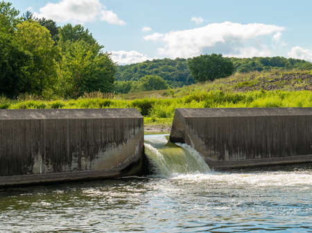 Water flows through the floodgate in a concrete dam on a river, with green trees and vegetation on...