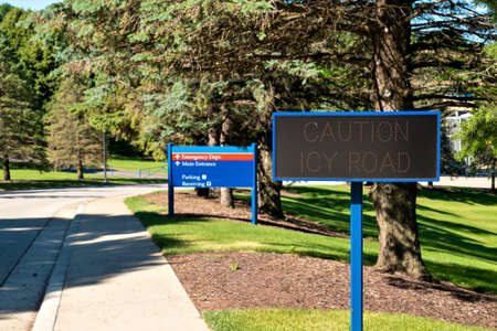 Electronic CAUTION ICY ROAD next to a street and sidewalk in summer, with green grass and evergreen trees, is ready for winter weather when it will be active when needed.の写真素材