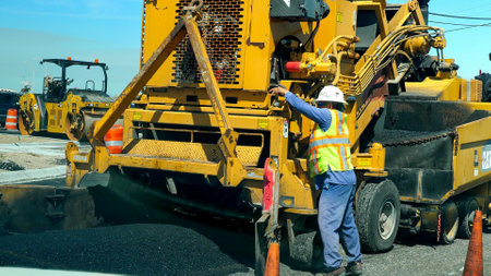 PORT ARANSAS, TX - 28 FEB 2020: Road construction machine laying new asphalt on city street. Working man in hardhat and yellow machine and fresh black asphalt tar, on a sunny day.のeditorial素材