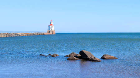 Lighthouse with red roof at the end of a pier on Lake Superior, and large boulders surrounded by ripples of blue water being blown away from the camera by the wind.の写真素材
