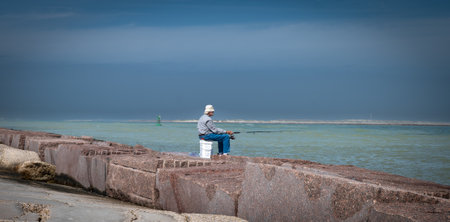 PORT ARANSAS, TX â 7 FEB 2023: Elderly fisherman sitting patiently on a plastic pail while fishing at the South Jetty in Port Aransas, Texas.のeditorial素材