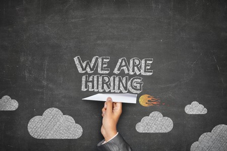 We are hiring concept on black blackboard with businessman hand holding paper planeの写真素材