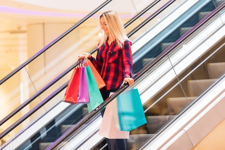 Young blonde smiling attractive woman with long hair with a lot of color shopping bags on escalator in the fashion storeの写真素材
