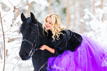 Young beautiful blonde woman wearing a long pink dress riding a black horse in the snowy winter forestの写真素材