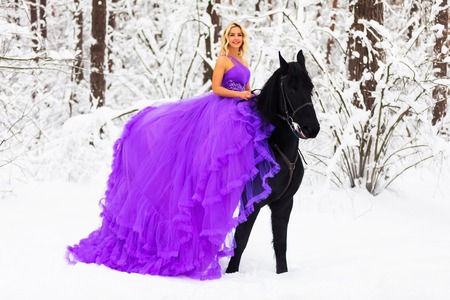 Young beautiful blonde woman wearing a long pink dress riding a black horse in the snowy winter forestの写真素材