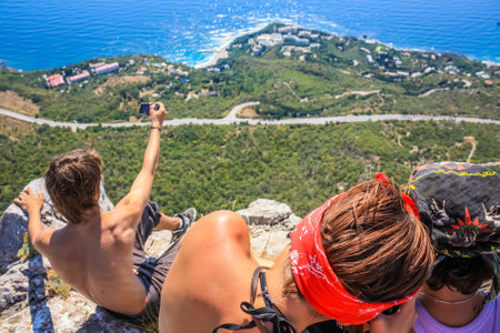 The girls and the guy are sitting on the edge of the mountain. Below you can see the blue sea. Man photographing. Crimeaの写真素材