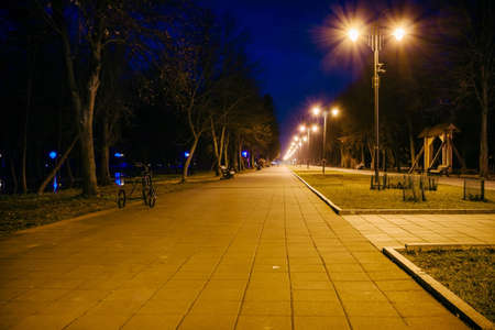 Night city park. Wooden benches, street lights and park alley. The tiled road in the night park with lanterns. Benches in the park at night. Illumination of a park road with lanterns at night. Lutskの写真素材