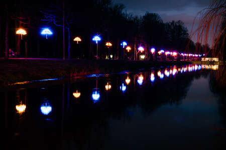 The road in the night park with illuminated umbrellas and the reflection in the river. Illumination of a park road with small lanterns on umbrellas suspended above the path at night. Lutsk.Ukraineの写真素材