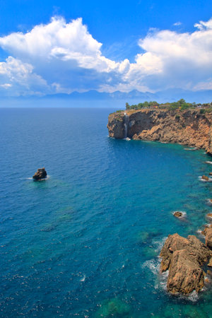 Stunning coastal landscape featuring a waterfall, rocky shore, and sunny skies with distant mountains in Antalya, Turkeyの写真素材