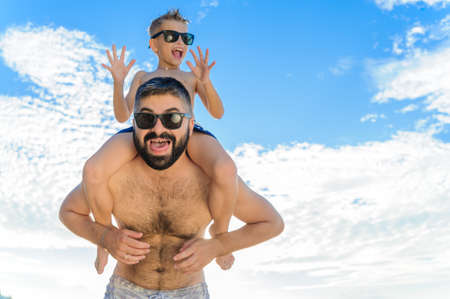 Eight years old boy sitting on dad's shoulders. Both in swimming shorts and sunglasses, having fun...