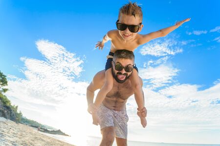 Eight years old boy sitting on dad's shoulders. Both in swimming shorts and sunglasses, having fun on the beach. Bottom view. Blue sky and altocumulus clouds behind themの写真素材