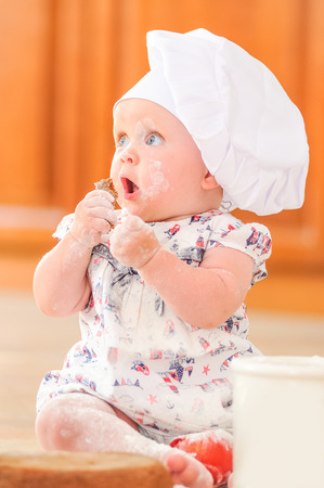 Cute liitle girl in chef's hat sitting on the kitchen floor soiled with flour, playing with food, making mess and having funの写真素材