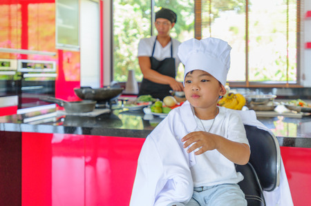 Cute little Thai boy in chef's hat and tunic sitting on a high chair in a modern style home kitchen. Dad is behind him making Thai foodの写真素材