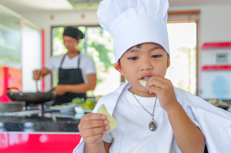 Cute little Thai boy in chef's hat and tunic sitting on a high chair in a modern style home kitchen. Dad is behind him making Thai foodの写真素材