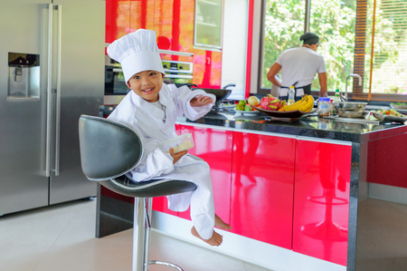 Cute little Thai boy in chef's hat and tunic sitting on a high chair in a modern style home kitchen. Dad is behind him making Thai foodの写真素材