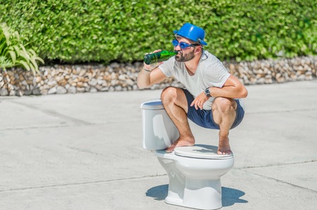 The bearded man in goggles astride the toilet, which is installed in the middle of the street. Toilet brush in his hand, cooking pan on his head. Drinking beerの写真素材