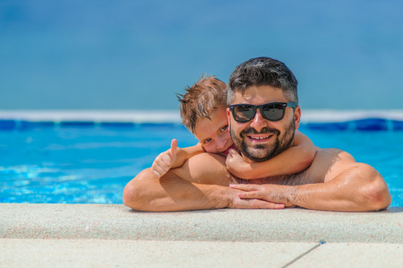 Seven years old cute boy smiling without tooth. He and his bearded father are in sunglasses, show their tongues, they leaned on pool railing giving a grimace of happiness. Sea view and beach umbrellasの写真素材