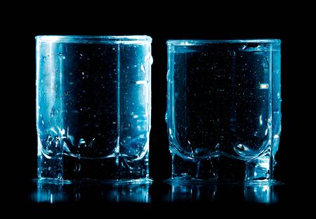 Splashes and drops of blue water in two glasses are isolated on a black background.の写真素材