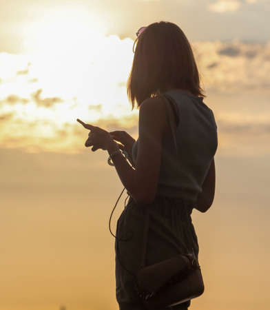 A girl from behind stands with a phone at sunset.のeditorial素材