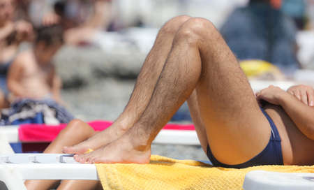 The legs of a man lies on a sun lounger on the beach by the sea.