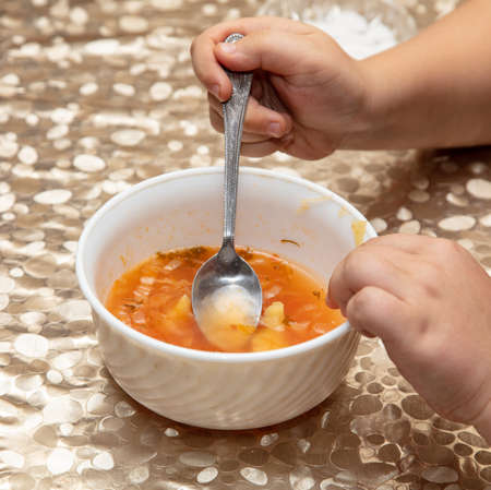 Boy eats soup with a spoon from a white plate.