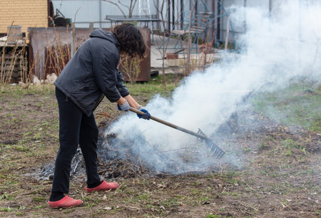 A young woman in a black jacket burns dry grass in the yard.の写真素材