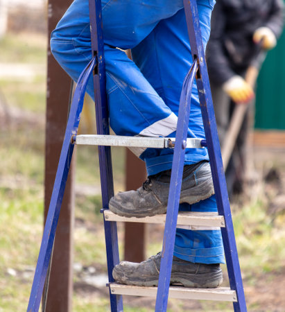 Worker in blue overalls stands on a metal ladder in the yard.の写真素材