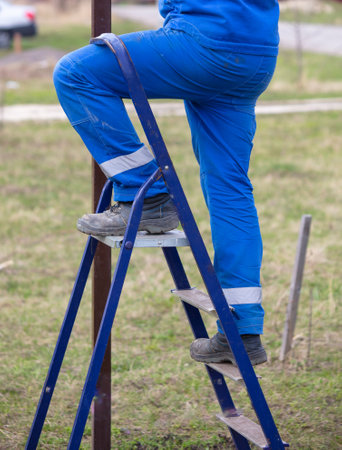 Worker in blue overalls stands on a metal ladder in the yard.の写真素材