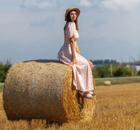 A girl in a dress and hat sits on a haystack.の写真素材