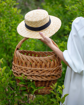 A woman is holding a basket with a straw hat on top of it. The basket is brown and woven. The woman is standing in a field of green bushesの写真素材