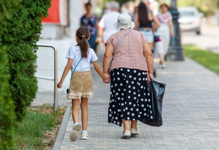 A woman with a child is walking down the street. The woman is wearing a polka dot skirt and a hat. The child is holding her hand. There are other people walking in the backgroundの写真素材