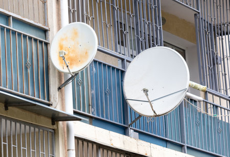 Two satellite dishes are mounted on a building. One is white and the other is brown. The brown dish is older and has a rusted appearanceの写真素材