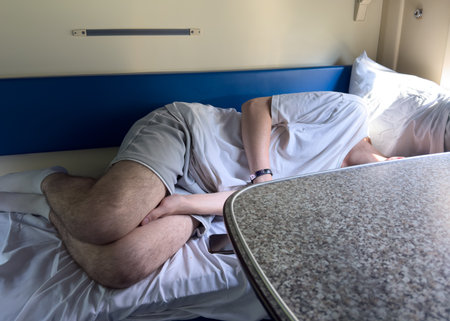 A man is sleeping on a bed with his head on a table. The bed is covered in white sheets and the table is made of woodの写真素材