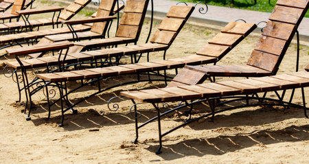 A row of wooden lounge chairs are set up on a sandy beach. The chairs are arranged in a line, with...